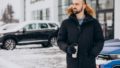 Handsome man in warm jacket standing by car covered with snow and drinking coffee