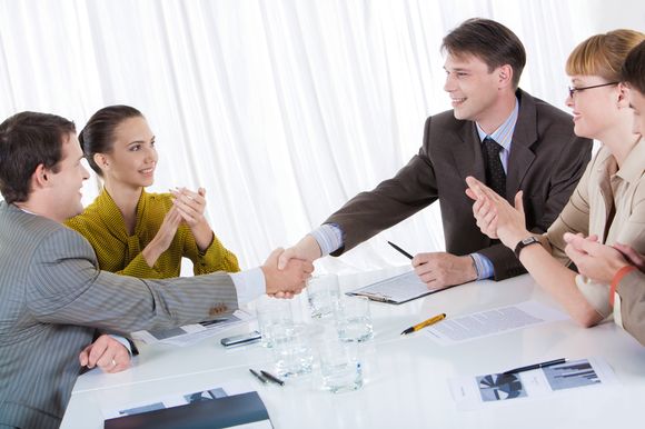 Business partners shaking hands after striking deal while their co-workers applauding