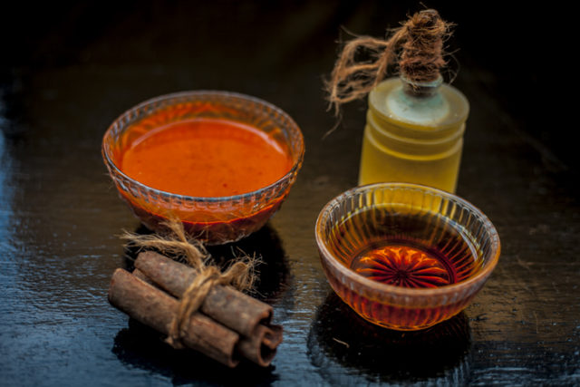 Cinnamon powder, honey and olive oil hair mask in a glass bowl on a wooden surface with gold lights isolated. Along with raw cinnamon stick, honey & olive oil present on the surface. Horizontal shot.