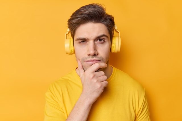 Horizontal shot of thoughtful man holds chin and looks seriously at camera listens favorite song via stereo headphones dressed in casual clothes poses against vivid yellow background. Let me think