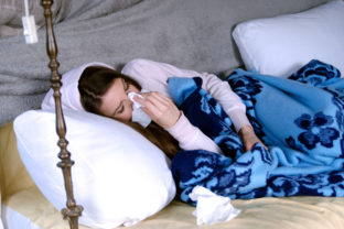 Sick woman with flu or alergy symptoms laying in bed under blanket,sneezing her nose in paper tissue