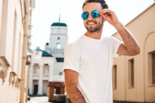 Portrait of handsome smiling stylish hipster lambersexual model.Man dressed in white T shirt. Fashion male posing on the street background in round sunglasses