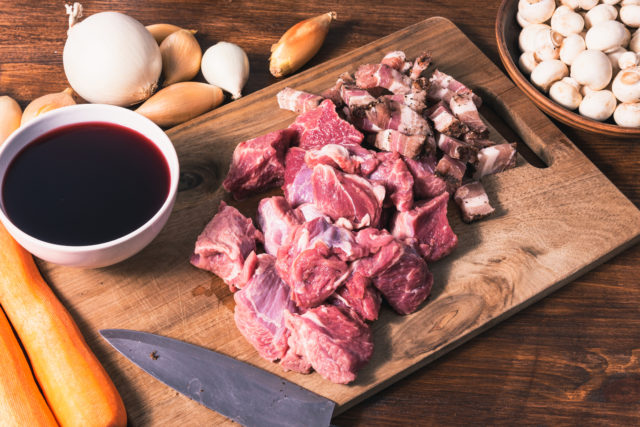 Ingredients for making burgundy beef: beef, shallots, carrots, bacon and red wine on a rustic style cutting board table.