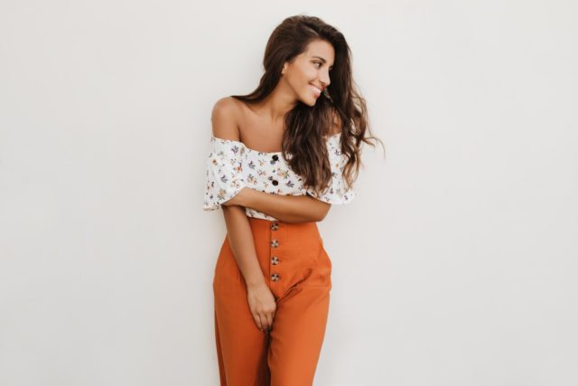 Picture of lady in orange trousers with high waist and white top with floral print. Brunette girl is cute smiling on white background.