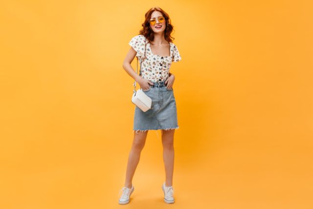 Cute redhead lady in skirt and stylish T shirt with smile posing on orange background.