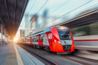 Electric passenger train drives at high speed among urban landscape.