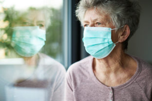 A old woman or grandma is wearing a respirator or surgical mask and looking out of the window