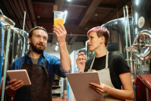 Young bearded brewer in apron looking at beer in glass while standing next to his female colleague
