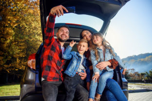 Happy stylish parents with their cute lovely children are making funny selfie on smart phone while sitting in the trunk. Happy modern family concept.