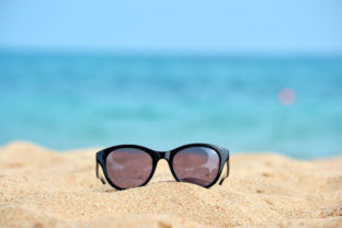 Closeup of black protective sunglasses on sandy beach at tropical seaside on warm sunny day. Summer vacation concept
